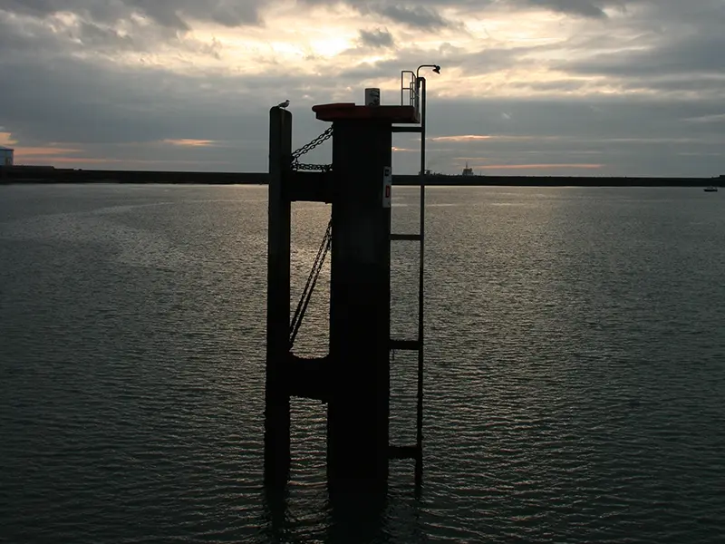 Une sorte de pylone de béton et de métal surgit au beau milieu de la mer et crée une masse sombre et géométrique qui contraste avec la trame des vaguelettes et de celle des nuages.